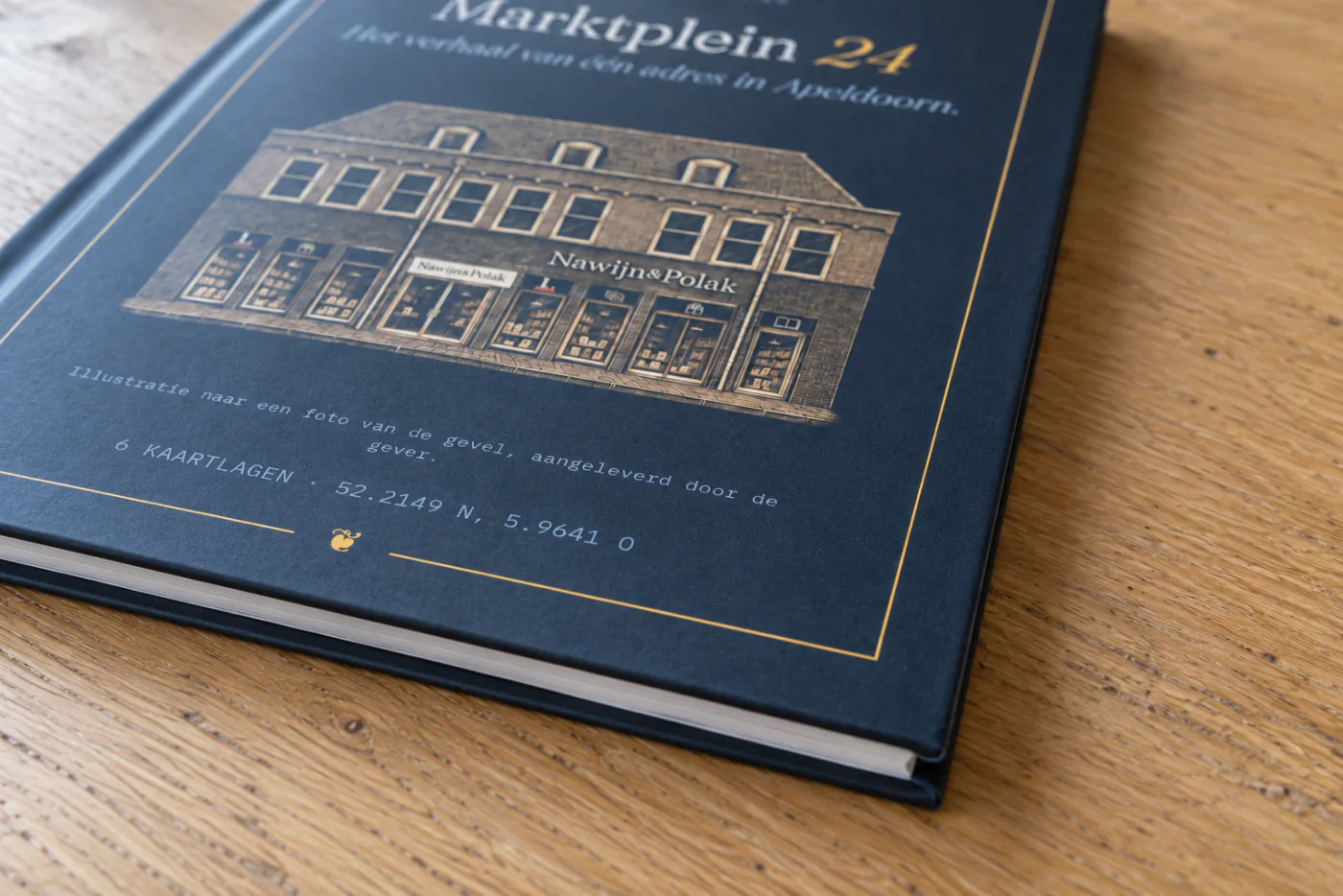 De kaft van dichtbij, schuin op een houten tafel: de donkerblauwe hardcover met gouden lijst, de getekende gevel en de witte rand van het papier
