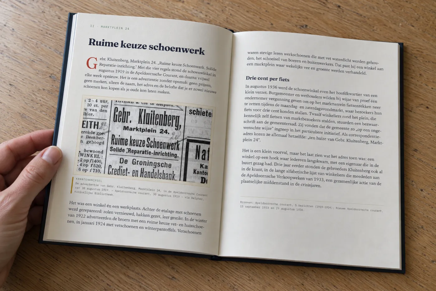 Het boek over Marktplein 24 opengeslagen op een houten tafel, een hand houdt de bladzijde vast: het hoofdstuk „Ruime keuze schoenwerk” met een krantenadvertentie van Gebr. Kluitenberg uit 1919 en de bron eronder