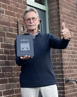 Een man houdt het gebonden boek naar de camera en steekt zijn duim op, voor zijn huis
