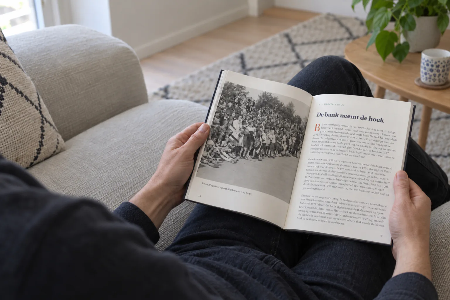 Iemand zit thuis op de bank en leest in het boek over Marktplein 24: links een oude foto van een menigte langs de straat, rechts het hoofdstuk „De bank neemt de hoek”